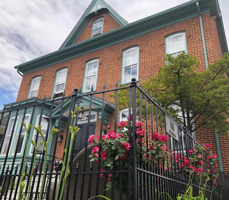 A two-story brick house with tall windows, teal trim, and a black metal fence. Pink flowers and greenery grow in front, and a tree stands by the entrance under a partly cloudy sky.