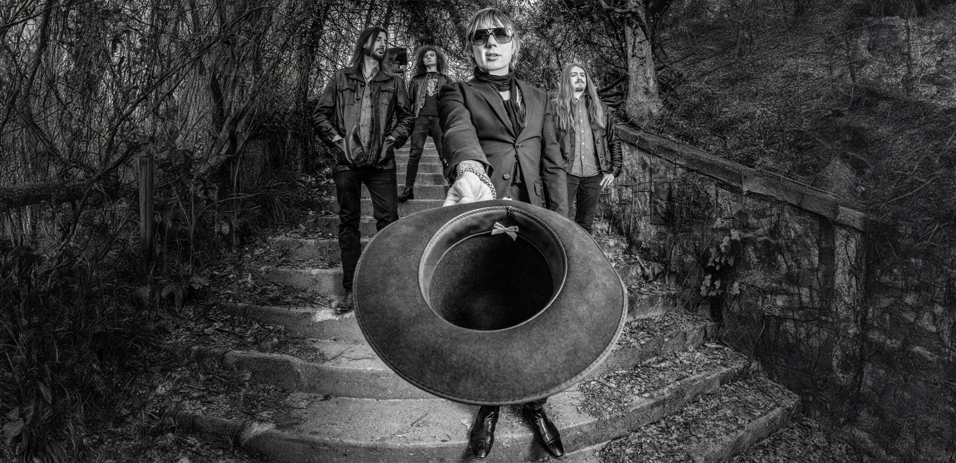 A black-and-white photo of five people standing outdoors on stone steps. The person in front, wearing sunglasses and a suit, extends a wide-brimmed hat toward the camera. Dense foliage surrounds the stone steps.