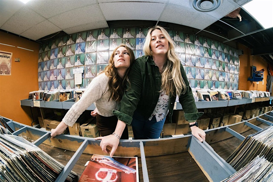 Two women smile and lean over bins of vinyl records in a record store, surrounded by colorful album covers displayed on the wall behind them. The photo has a wide-angle, slightly distorted perspective.
