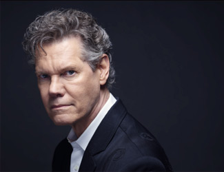 A man with curly gray hair, wearing a black suit jacket and white shirt, looks seriously into the camera against a dark background.