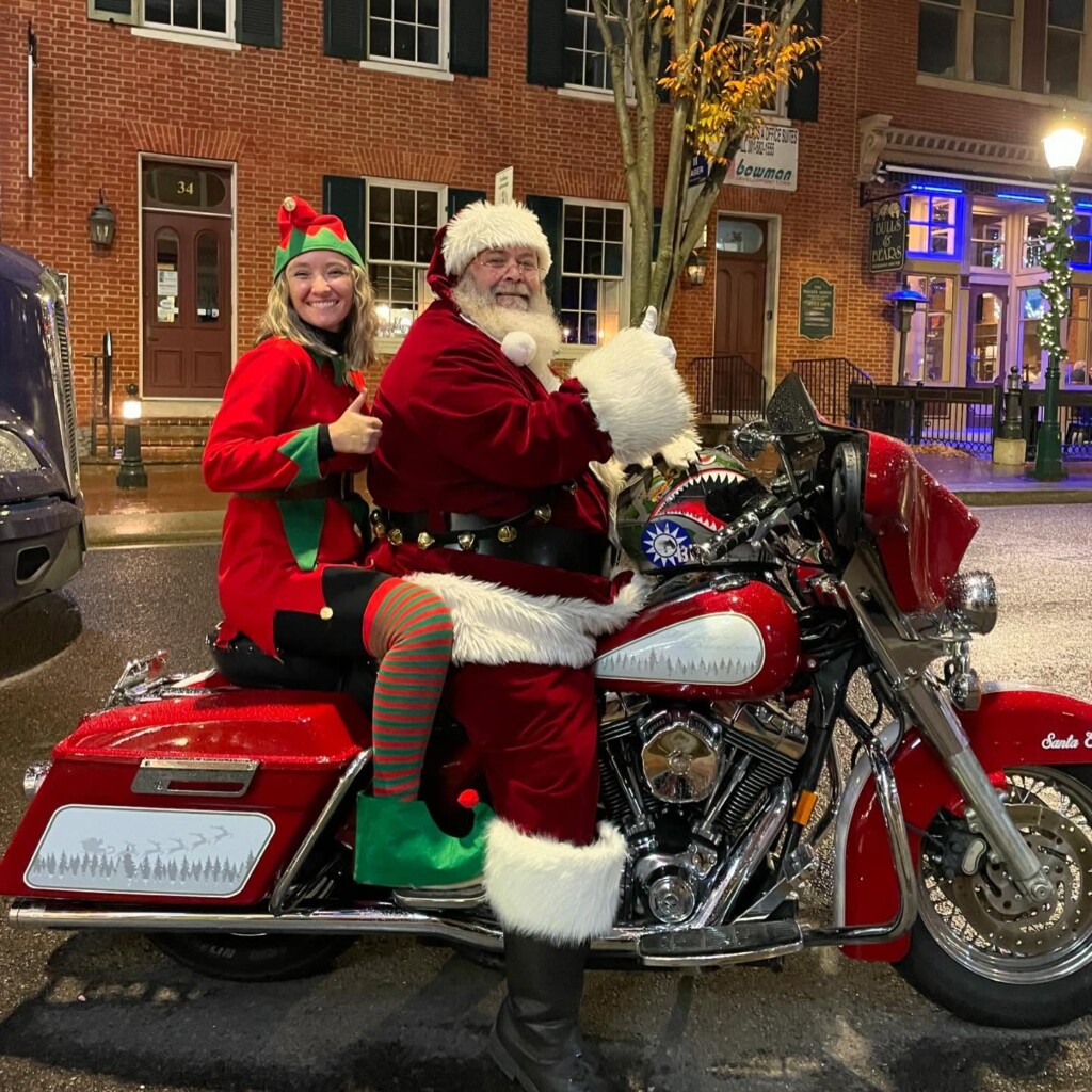 Santa Claus and a woman dressed as an elf sit smiling on a shiny red motorcycle decorated for the holiday season, parked on a wet street in front of brick buildings adorned with festive holiday lights.