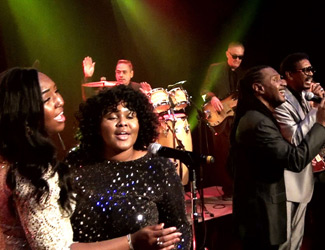 A lively band performs on stage under colorful lights, featuring two women singing in the foreground and four men playing instruments and singing in the background.