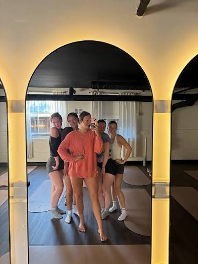 Five people pose for a group selfie in front of a large mirrored wall in a studio, standing close together and smiling, with warm lighting creating a cozy atmosphere—each showing off their unique style as if they were ready for an outfitters photoshoot.