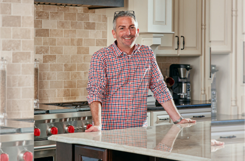 A man wearing a red and blue checkered shirt smiles while standing in a modern kitchen, resting his hands on a marble countertop. The kitchen features light cabinetry, a coffee maker, and a tiled backsplash.