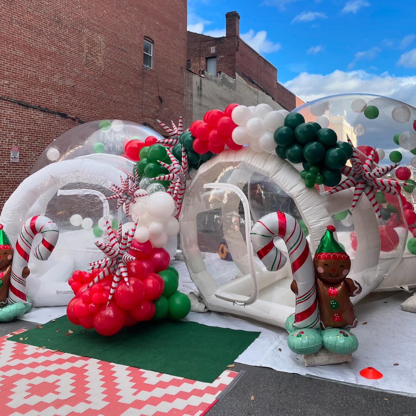 Two clear inflatable domes decorated with red, white, and green balloons, large candy canes, pinwheels, and gingerbread figures create a festive holiday-market scene outdoors against brick buildings under a blue sky with clouds.