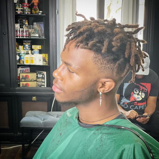 A young man with short, twisted dreadlocks and a fresh fade haircut sits in a barber chair, wearing a green cape. He has a cross earring and a thin chain necklace. Another person sits in the background.