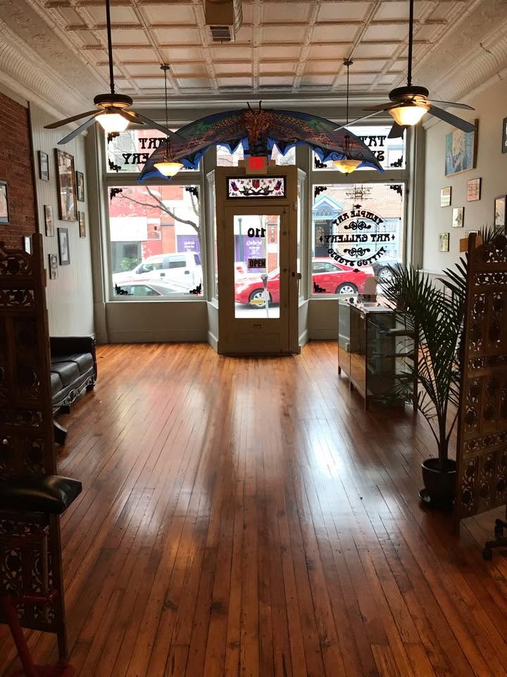 A welcoming tattoo studio with wooden floors, decorative chairs, a cozy sofa, artwork on the walls, and large windows facing the street where cars can be seen outside.