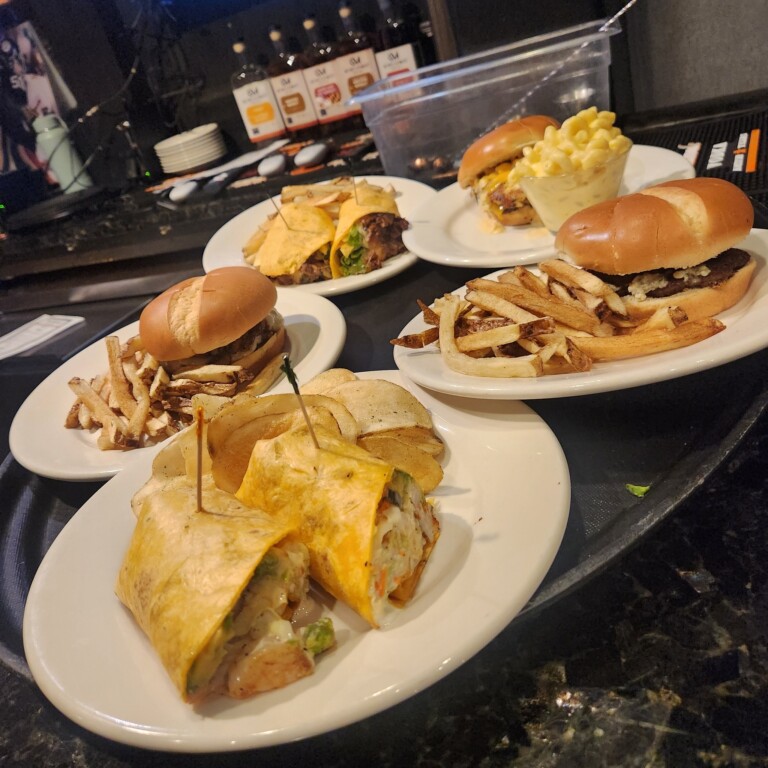 Three plates with burgers and fries, one plate with a wrap cut in half, and one plate with a burger and macaroni and cheese, all arranged on a tray on a dark countertop at a restaurant.