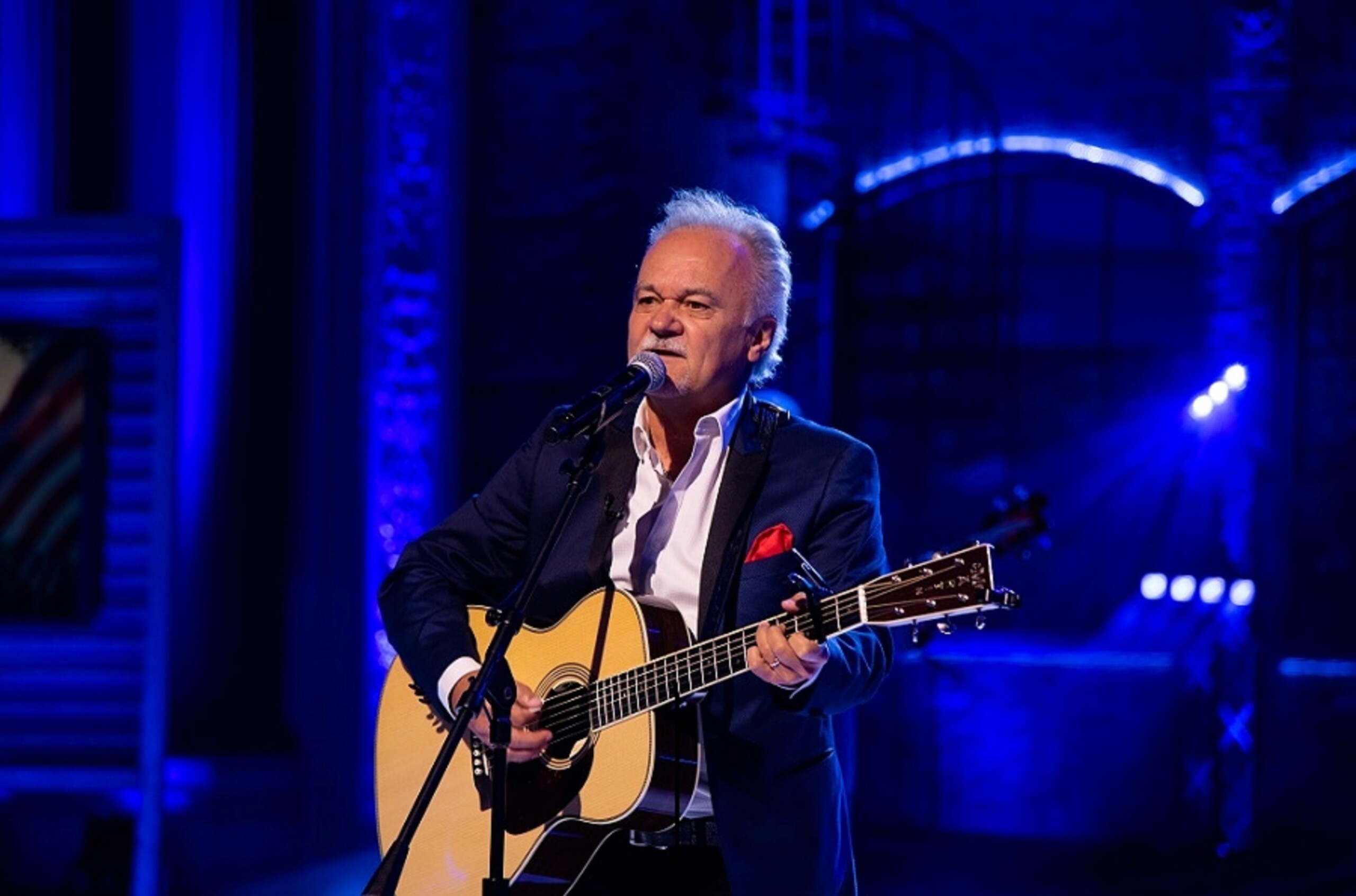 An older man in a dark suit plays an acoustic guitar and sings into a microphone on a blue-lit stage.