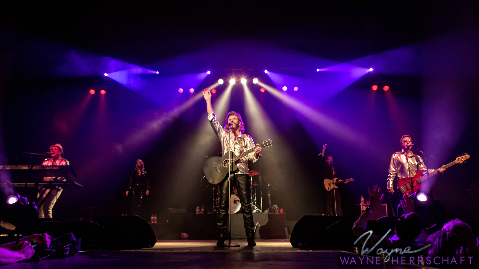 A band performs on stage under colorful spotlights. The lead singer stands center stage with a guitar, one arm raised. Other band members play keyboard, guitar, and bass. Audience hands are visible, and the stage is vibrant and lively.