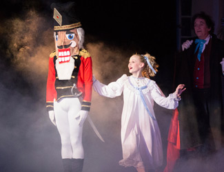 A young girl in a white nightgown smiles and touches the arm of a person dressed as a Nutcracker soldier on stage, surrounded by theatrical fog, with another person watching in the background.