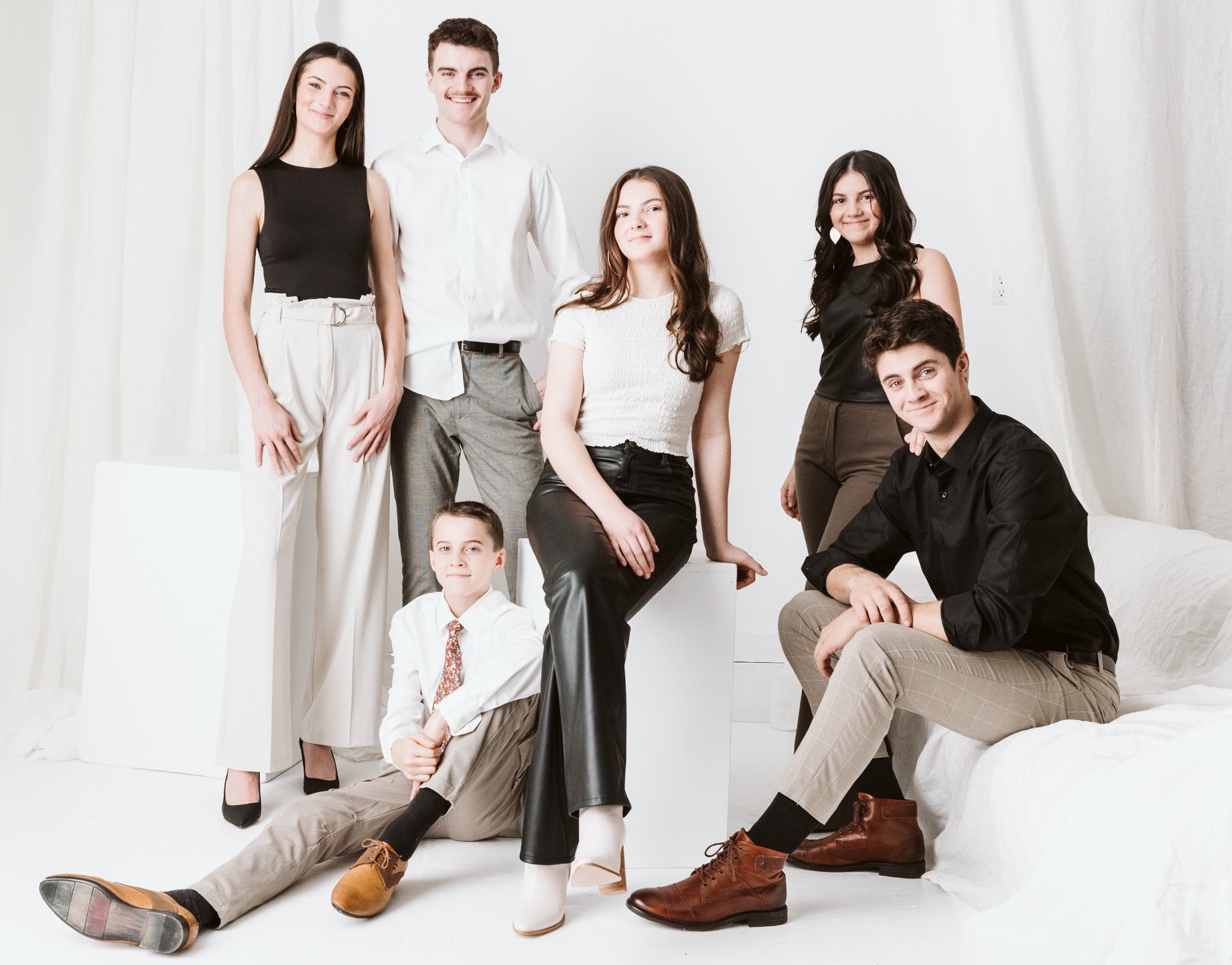 Six young people pose together in a bright, minimalist room. Three stand, two sit on cubes, and one sits on the floor. They wear smart-casual outfits in neutral tones, and all are smiling at the camera.