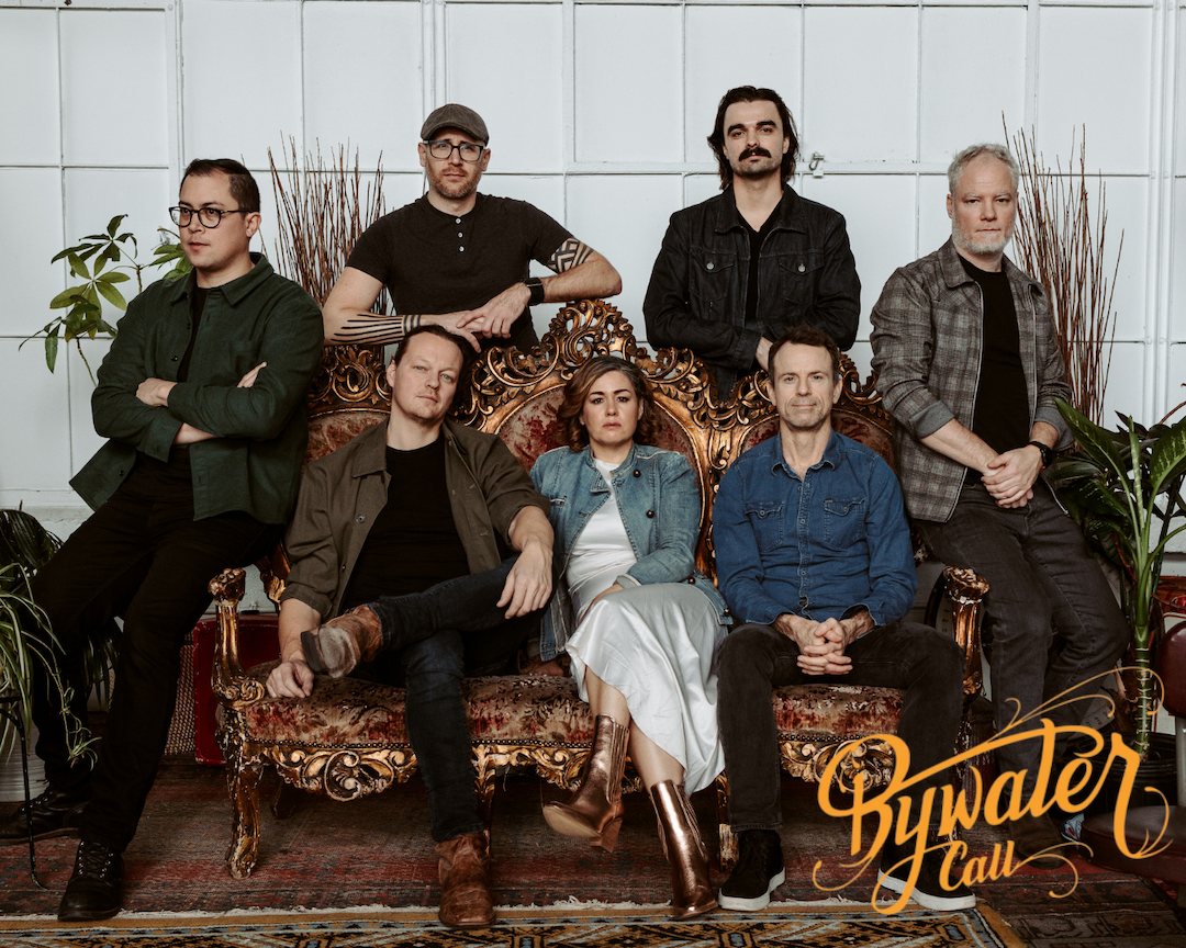 Seven people pose together indoors, seated and standing around an ornate sofa with gold accents. They have neutral expressions. The "Bywater Call" logo appears in the lower right corner.