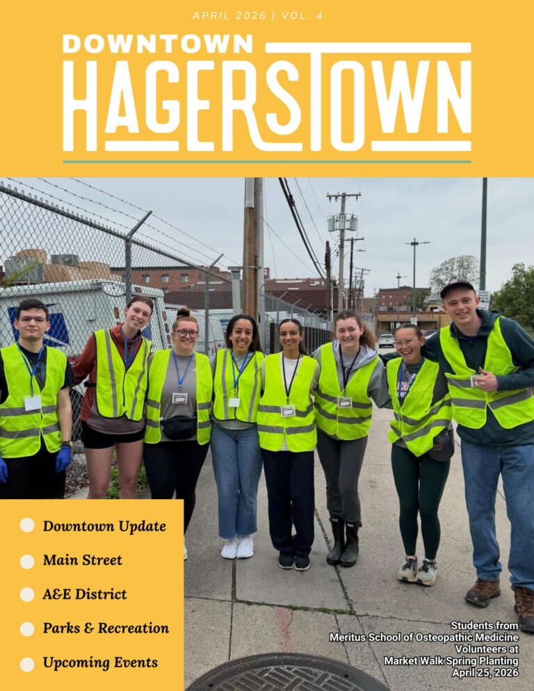 A group of nine volunteers in yellow safety vests pose and smile for a photo on a sidewalk, with a cityscape and chain-link fence in the background. Text highlights Downtown Hagerstown newsletter updates and upcoming events.