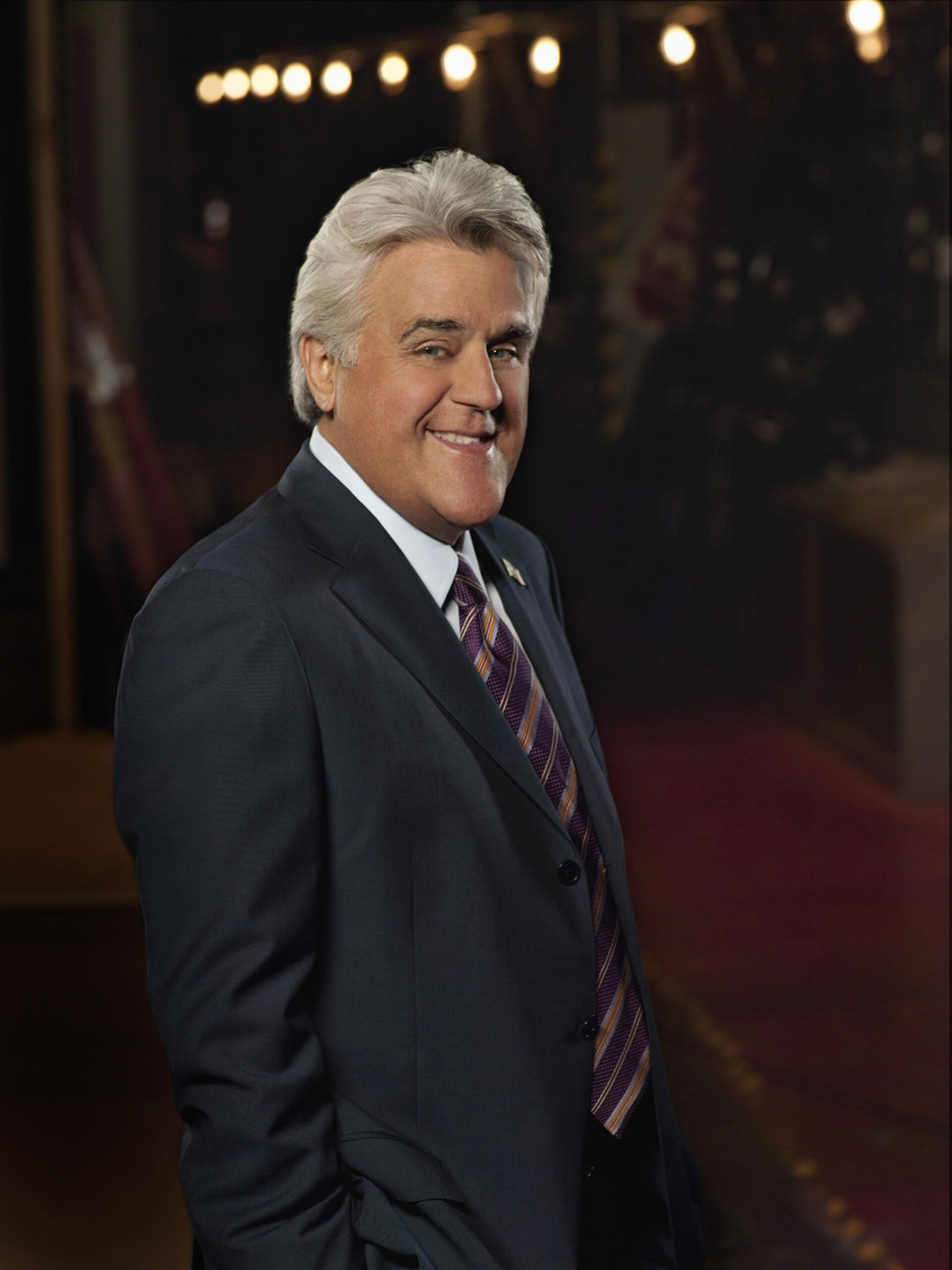 A man with gray hair wearing a dark suit, striped tie, and white shirt stands smiling in front of a dimly lit background with string lights.