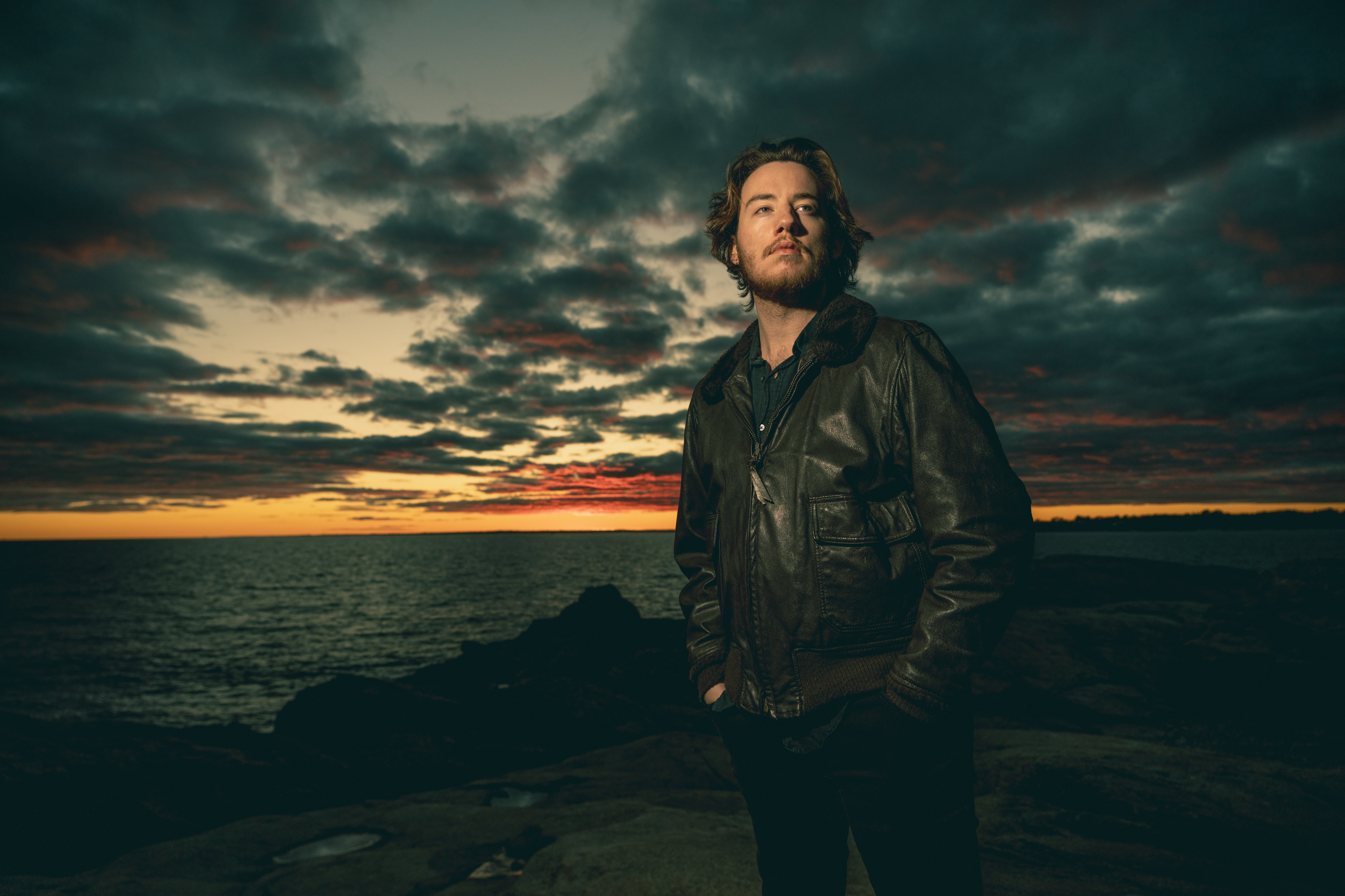 A man in a dark leather jacket stands with hands in his pockets on rocky ground by the sea at sunset, with dramatic clouds and orange hues in the sky behind him.
