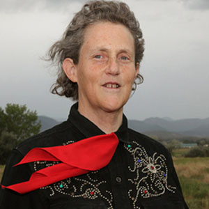 A person with short curly gray hair stands outdoors, wearing a black shirt with embroidered designs and a red scarf. The background shows trees, grass, and mountains under a cloudy sky.