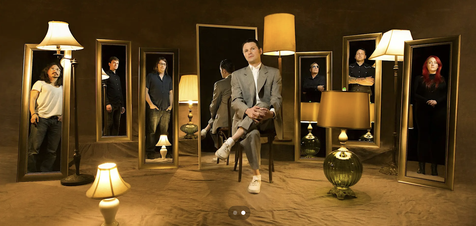 A person in a gray suit sits on a chair, surrounded by floor and table lamps and seven people reflected in tall mirrors on a dark brown background.