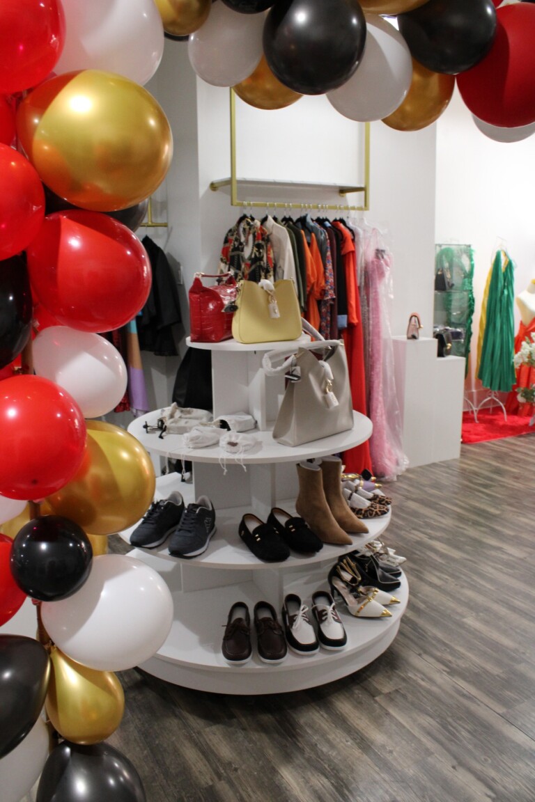 A clothing boutique interior with a round display table holding shoes, boots, handbags, and accessories, decorated with a colorful balloon arch in red, gold, black, and white. Clothes hang on racks in the background.
