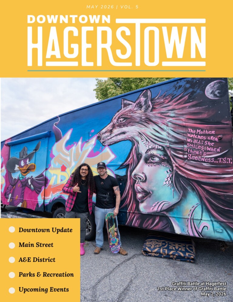 Two people pose in front of a mural-covered van featuring a wolf and a woman. The image, also used in the “Downtown Hagerstown” newsletter, highlights local updates and events on the magazine cover.