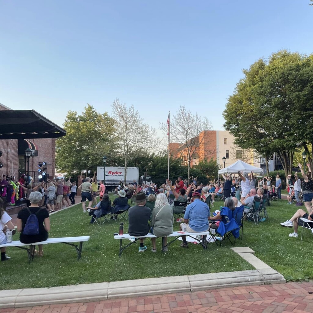 A crowd of people sits on folding chairs and benches on a grassy lawn, watching a live outdoor performance near a stage on a sunny day. Trees and buildings surround the park area.