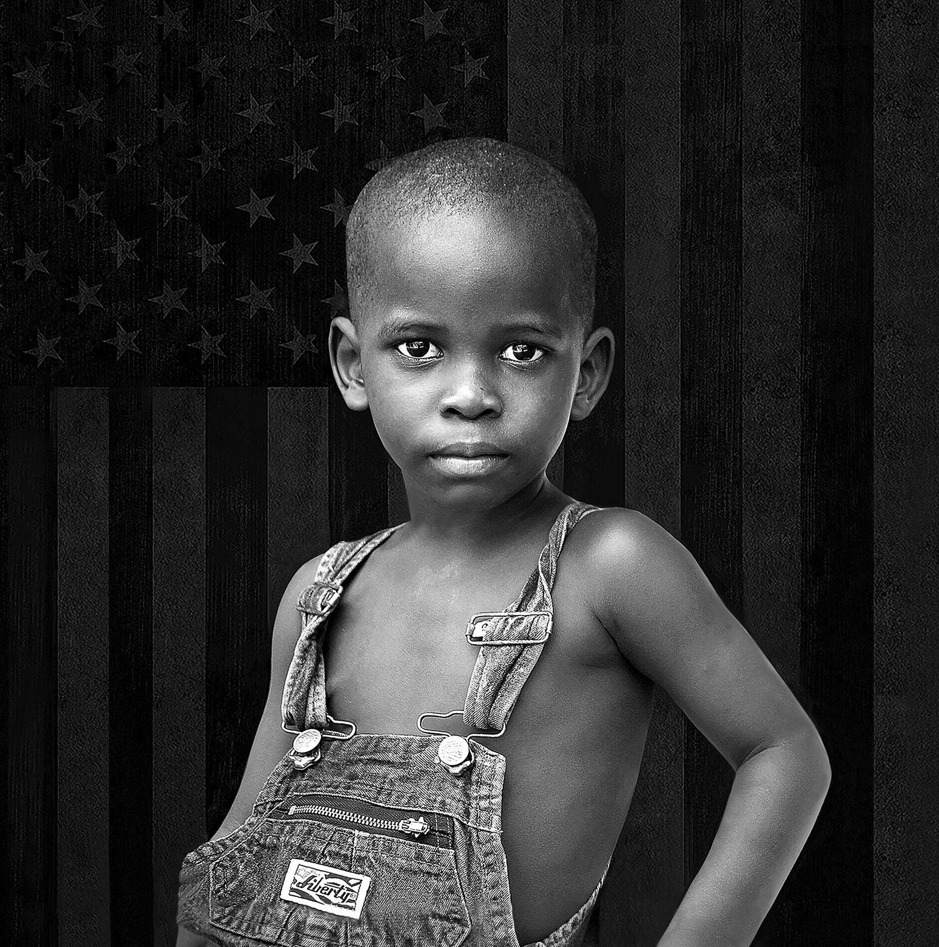 A young, black child wearing denim overalls stands confidently with one hand on their hip, gazing at the camera. A faded, monochrome American flag is visible in the background.