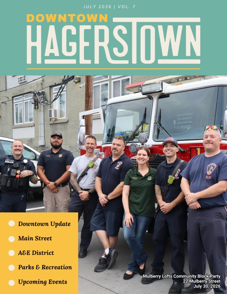 A group of men and women, some in uniform, stand smiling in front of a fire truck outside a building. A newsletter header reads “Downtown Hagerstown,” with event and feature highlights listed on the lower left.