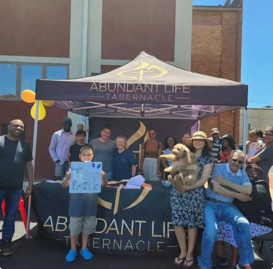 A group of people, including children and a dog, smile and pose under an "Abundant Life Tabernacle" tent at an outdoor event. Some hold signs, and balloons are attached to the tent in the background.