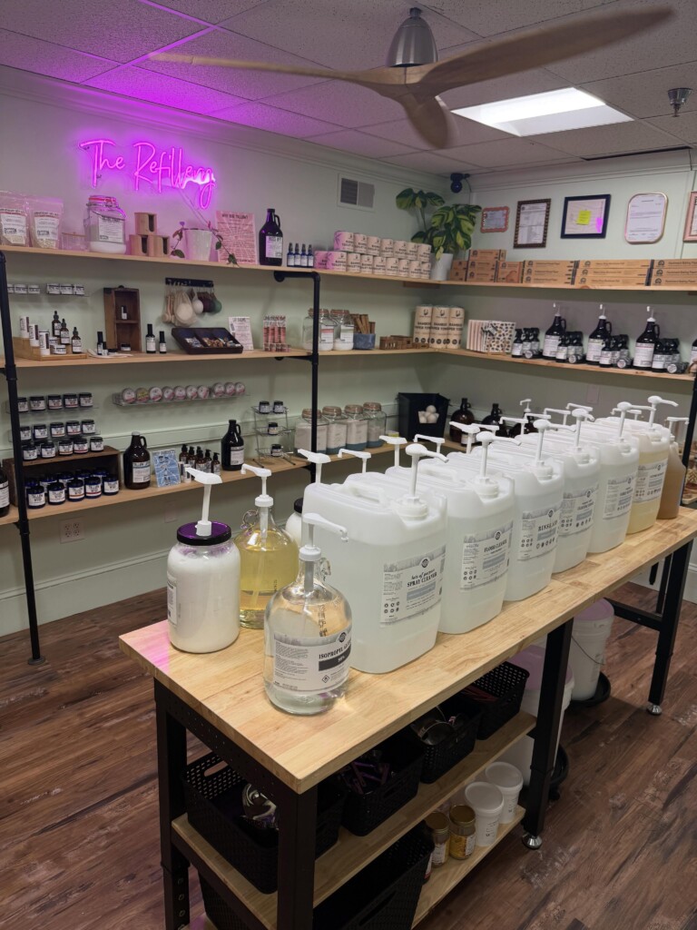 A sage green room rounded with bamboo shelves and black steel pipes as the base for the shelves. The shelves hold glass products like Tallow Whip, Soap. bamboo paper towels, soap, and more. A bammboo table with black legs stands in the middle of the room and holds an assortment of cleaner tubs that patrons can use to refill bottles.