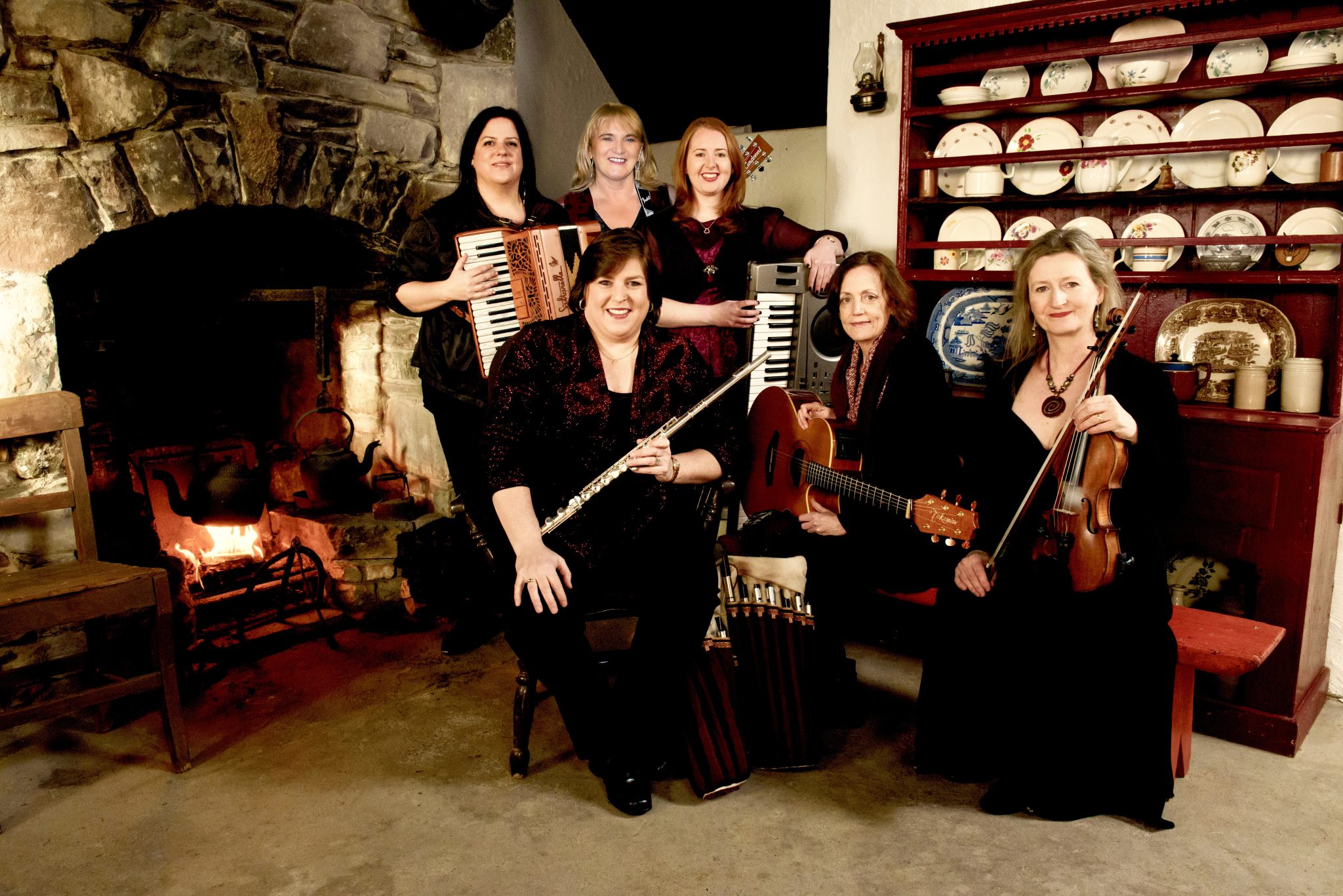 Six women with musical instruments, including a flute, violin, guitars, and keyboards, pose together in a cozy room with a stone fireplace and a red cabinet filled with dishes. All are smiling.