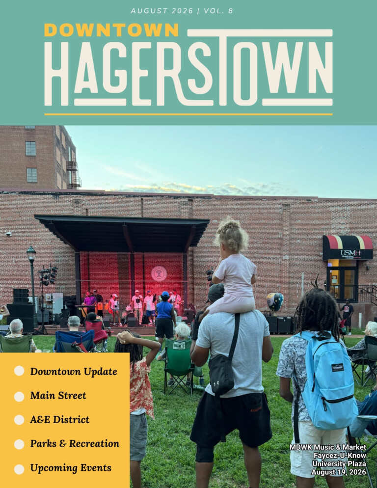 People watch a live outdoor band in a park surrounded by buildings. A man carries a child on his shoulders. Newsletter details updates and upcoming events in Hagerstown are displayed in bright yellow boxes.