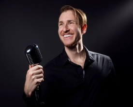 A smiling man in a black shirt holds a vintage microphone against a dark background, looking off to the side.