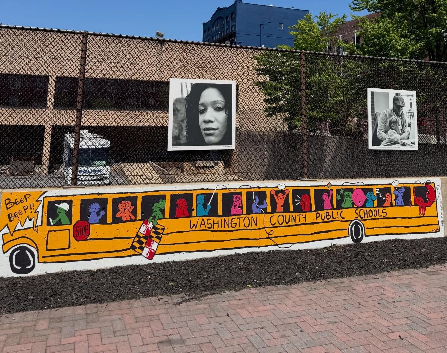A mural of a yellow school bus labeled “Washington County Public Schools” is painted on a low wall, with black-and-white portraits and a chain-link fence in the background, under a clear blue sky—a vivid reminder of the vibrant communities we create together.