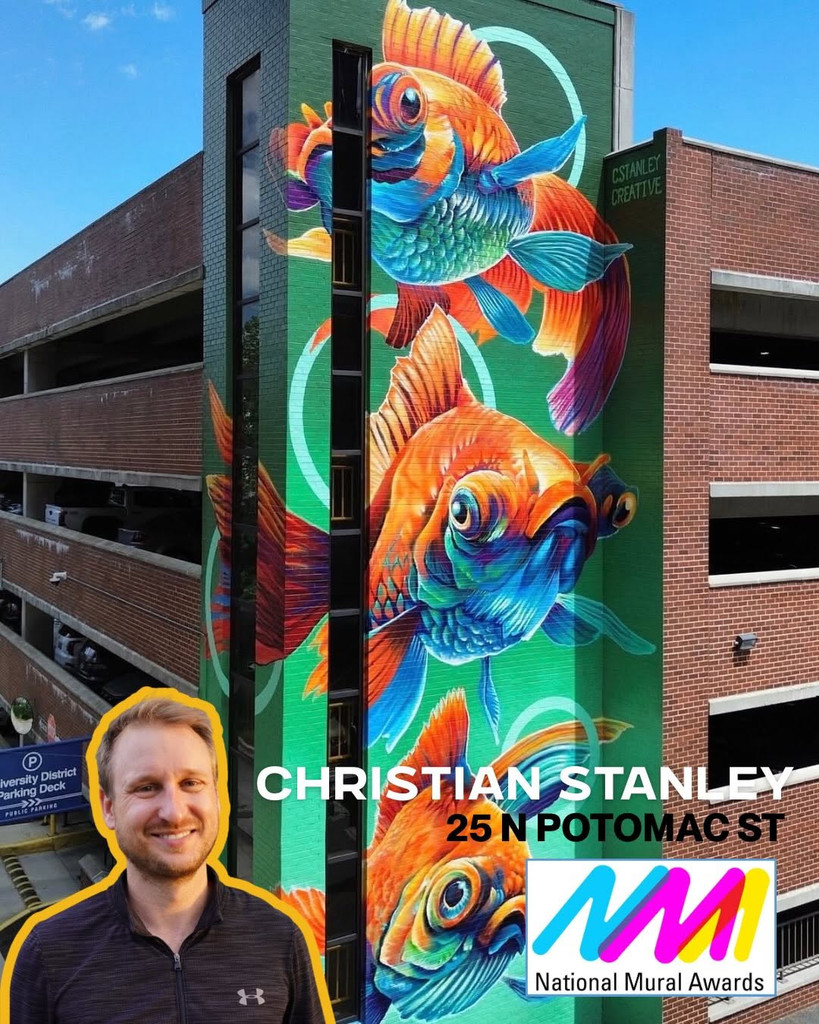 A tall mural of vibrant orange and blue fish brings life to the side of a building. A smiling man stands in the foreground, highlighting the striking mural’s presence. Text reads “Christian Stanley, 25 N Potomac St” with the National Mural Awards logo at the bottom right.