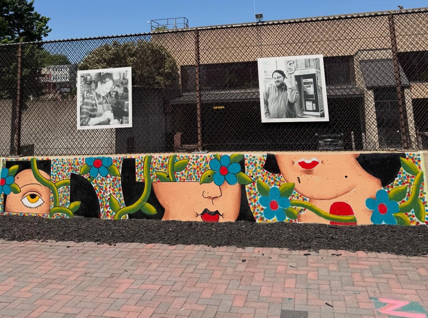 Two black-and-white photos hang on a fence above a colorful mural with faces and flowers—a vibrant testament to the art we create—set against a clear blue sky and a tan building in the background.