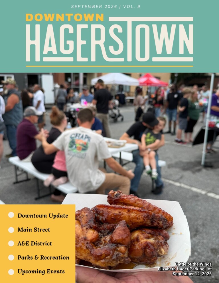 A plate of saucy chicken wings is held up at an outdoor food festival, with people mingling at tents and tables in the blurred background. The cover highlights events and updates for Downtown Hagerstown in this vibrant newsletter.