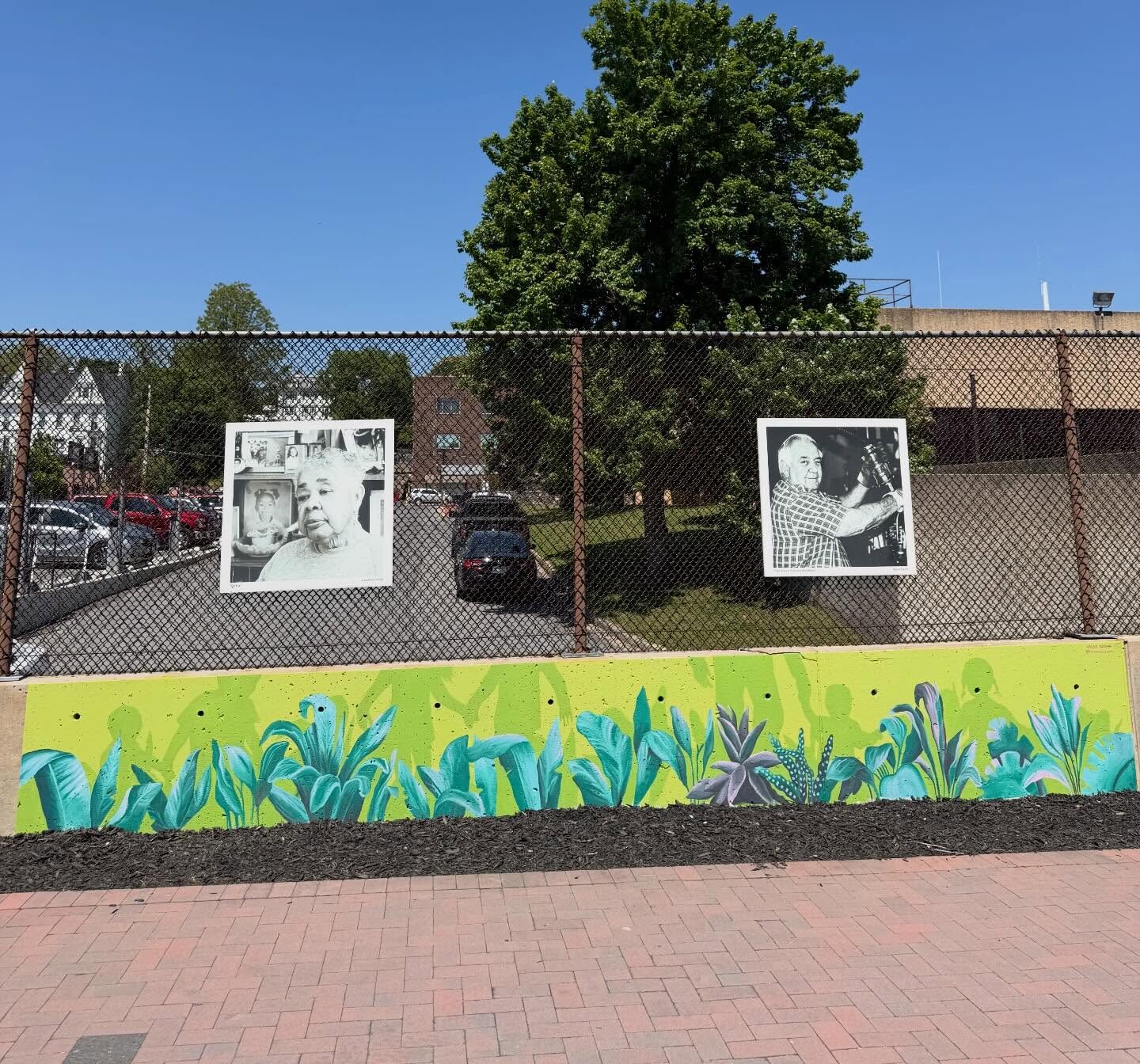 Photos of two people are displayed on a chain-link fence above a colorful mural with green plants, reflecting the vibrancy we create in our community, with a tree, parking lot, and buildings in the background under a clear blue sky.