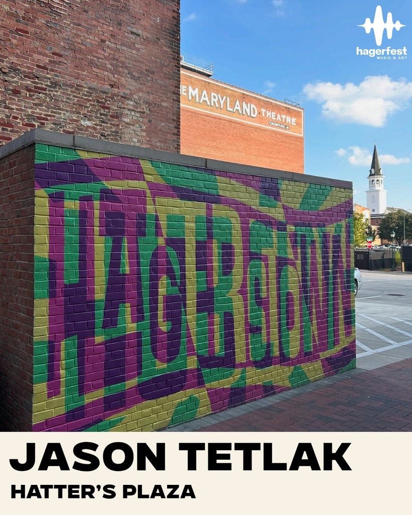 Colorful mural reading “HAGERSTOWN” on a brick wall in a plaza, with the Maryland Theatre and a church steeple in the background. Text at the bottom reads: “Jason Tetlak HATTER’S PLAZA.”.