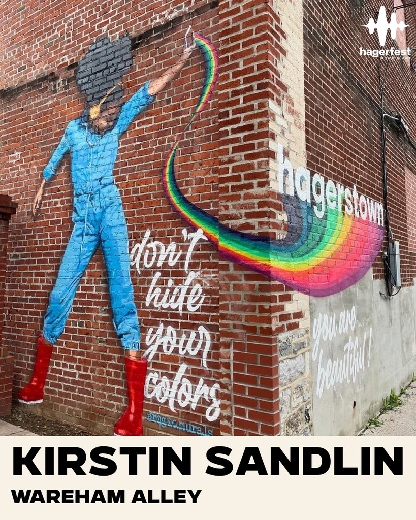 A mural of a woman in blue, red boots, raising her fist, with a rainbow and the words “don’t hide your colors”, “hagerstown”, and “you are beautiful!” painted on a brick wall. Text: Kirstin Sandlin, Wareham Alley.