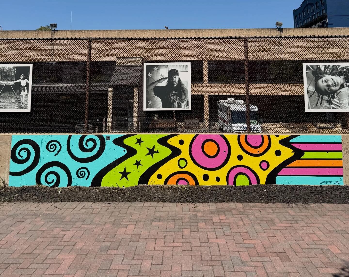 A colorful mural with swirls and dots—showcasing how we create vibrant public art—is in front of a fence, with black-and-white portrait photos displayed on the building behind it under a clear blue sky.