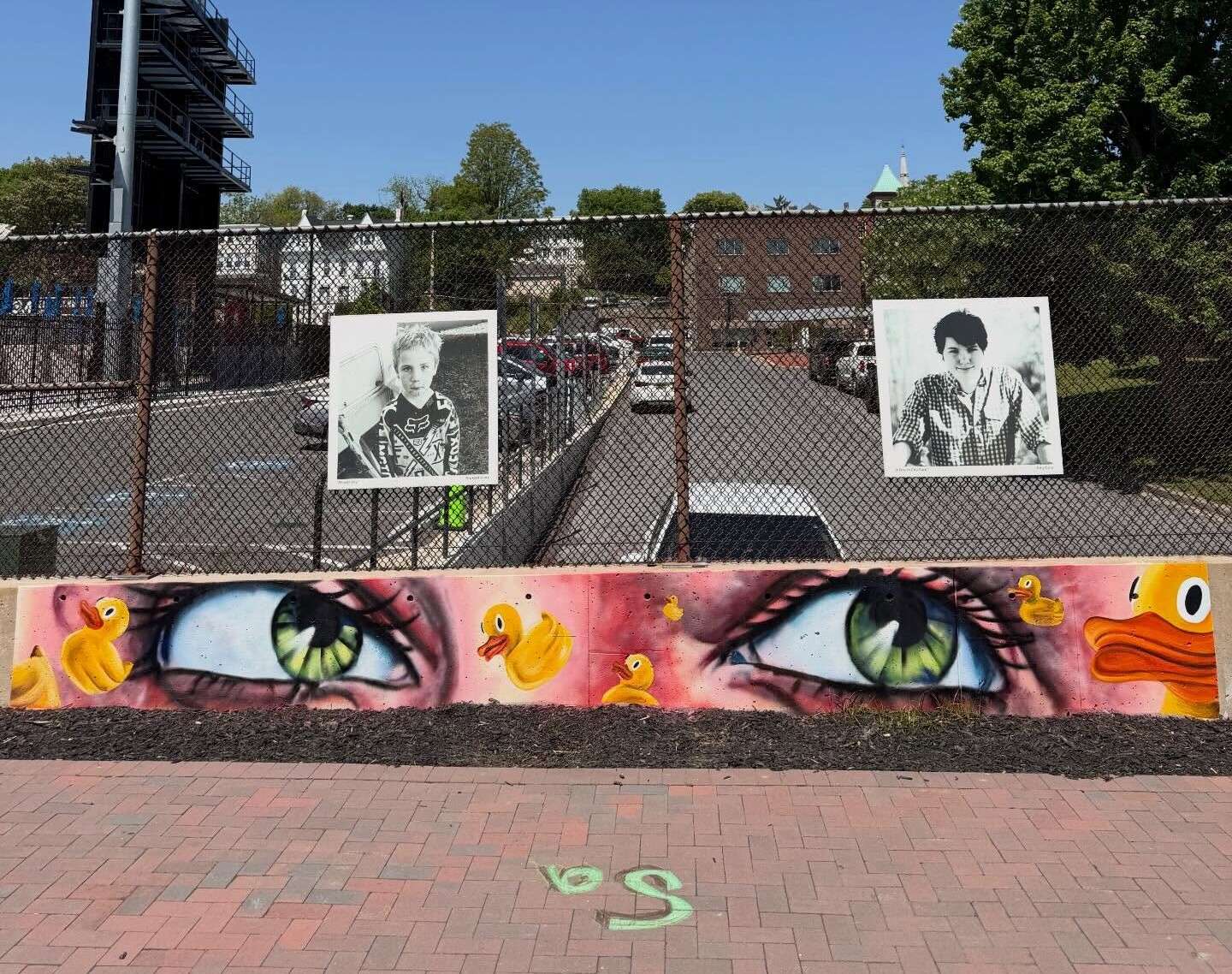 A mural of wide eyes with rubber ducks is painted on a low wall, a playful reminder of the unique art we create. Behind it, black-and-white portraits hang on a fence above a parking lot under a clear blue sky. Trees and a tall light pole are in the background.