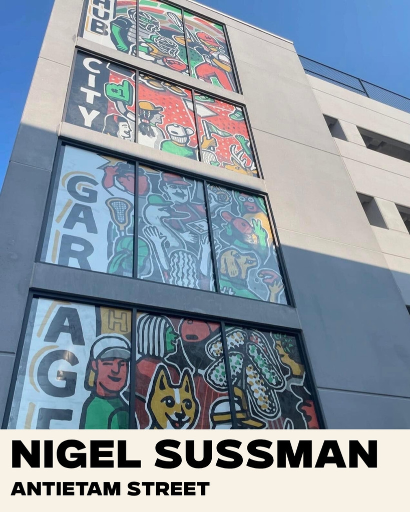 Colorful mural with cartoon figures, a dog, and vibrant patterns decorates several windows of a parking garage. Text reads “CITY GARAGE.” A label at the bottom says “NIGEL SUSSMAN, ANTIETAM STREET.”.