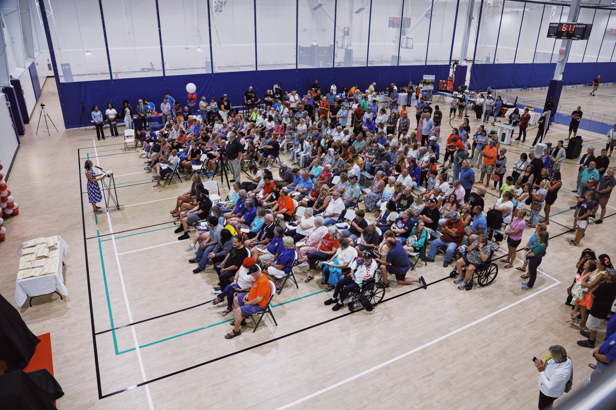 A large group of people, some seated and some standing, attend an indoor event in a gymnasium. A speaker stands at the front near a podium, while attendees watch and listen—many holding copies of the event newsletter.