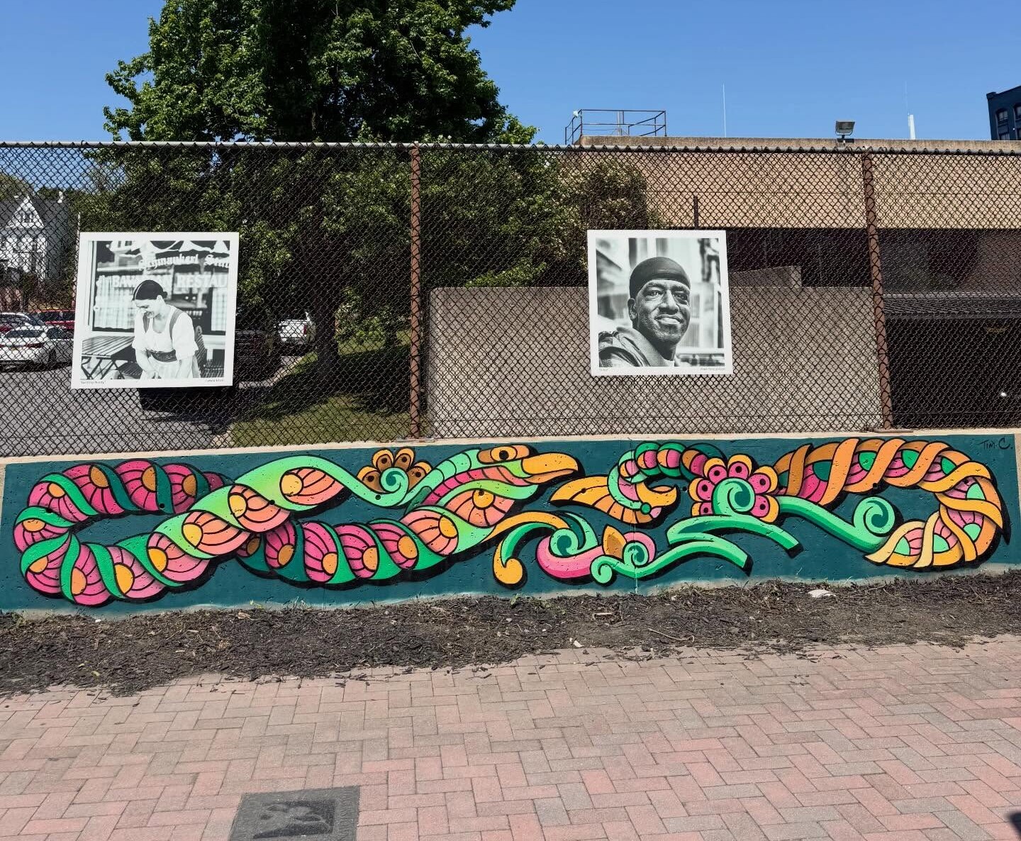A colorful mural with swirling patterns, which we create with passion and artistry, is painted on a low wall below two black-and-white portraits, set against a fence with a tree and building in the background.
