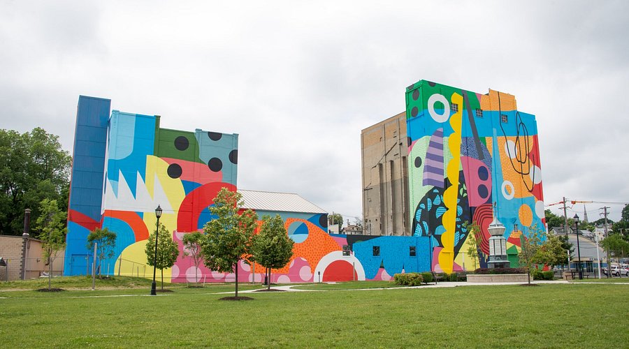 A vibrant abstract mural featuring geometric shapes and patterns covers tall building facades, set in a green park with trees, winding paths, and a small fountain under a cloudy sky.